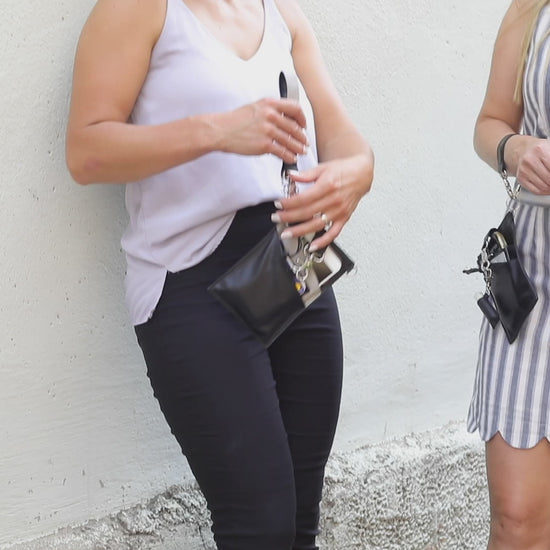 A person with a  black vegan leather phone pouch with a black vegan leather magnetic wristlet key chain and a black removable three pocket wallet and black lip balm holder set. Another Person  with a black and gray vegan leather bracelet, keychain, three pocket wallet, and a black lip balm holder used as a fanny pack belt bag. 